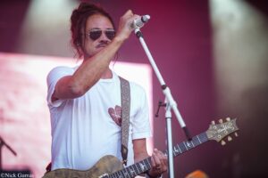 Male guitarist wearing sunglasses and a white tee, singing into a microphone while playing a wood-grain electric guitar on stage.