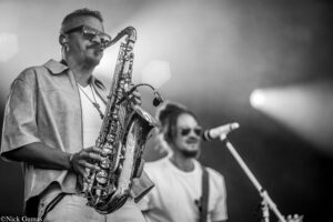 Saxophonist in sunglasses playing a horn on stage, with a singer at a microphone in the background (black-and-white).