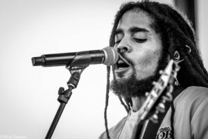 Black-and-white close-up of a man singing into a microphone, with dreadlocks visible nearby.