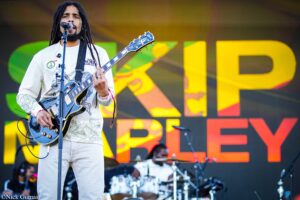 Male guitarist with dreadlocks singing into a microphone while playing a blue electric guitar on stage with a colorful backdrop behind him.