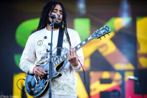 Musician with dreadlocks singing into a microphone while playing a blue electric guitar on a colorful outdoor stage.