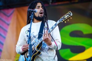 Guitarist with dreadlocks singing into a microphone on an outdoor stage, playing a blue electric guitar on a bright backdrop.