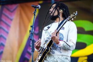 Musician with dreadlocks playing an electric guitar on stage, singing into a microphone.