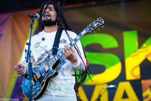 Musician with dreadlocks singing into a microphone while playing a blue electric guitar on a colorful stage backdrop.