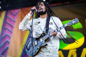 Musician with long dreadlocks singing into a microphone while playing a black electric guitar on stage.
