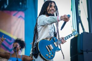 Male guitarist with long dreadlocks playing a blue electric guitar on stage, smiling at the crowd