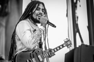 Male musician with long dreadlocks singing into a handheld microphone while playing an electric guitar on stage, in black and white.