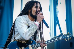 Guitarist with long dreadlocks singing into a handheld microphone on stage, wearing a light shirt and playing a black electric guitar.