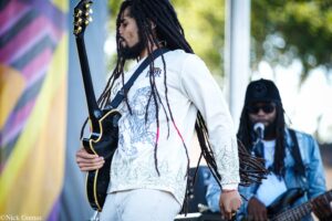 Musician with long dreadlocks in a white printed jacket plays an electric guitar on an outdoor stage, another guitarist in background wearing sunglasses and a cap.