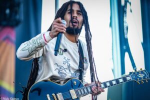Male guitarist with long dreadlocks sings into a microphone while playing a blue electric guitar on stage.
