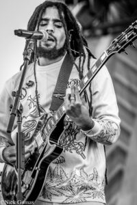 Black and white photo of a male guitarist with dreadlocks singing into a microphone while playing an electric guitar on stage