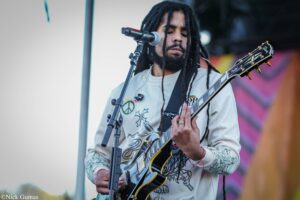 Musician with dreadlocks playing an electric guitar on stage, singing into a microphone nearby.
