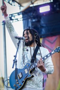 Male musician with dreadlocks singing into a microphone while playing a blue electric guitar on stage, one arm raised high in the air.