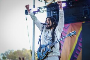Guitarist with dreadlocks on an outdoor stage, arms raised in the air while singing into a microphone.