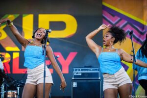 Two female singers perform on a colorful outdoor stage, microphones ready and arms raised in sync.