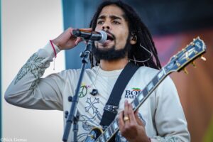 Male singer with dreadlocks and beard, wearing a light sweater, passionately singing into a microphone while playing an electric guitar on stage.