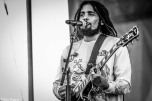 Black-and-white image of a man with dreadlocks singing into a microphone while playing an electric guitar on stage.