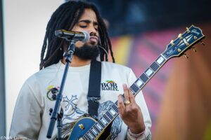 Male musician with dreadlocks sings into a microphone while playing a blue Gibson electric guitar on stage.