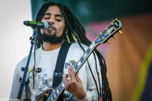 Musician with long dreadlocks singing into a microphone while playing an electric guitar on stage during a live performance