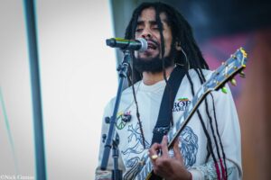 Male musician with long dreadlocks singing into a microphone while playing an electric guitar on a stage.