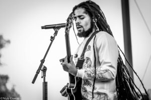 Guitarist with long dreadlocks playing an electric guitar and singing into a microphone on stage (black-and-white).