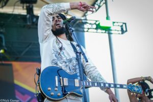 Male guitarist and vocalist performing on stage, singing into a microphone with a blue electric guitar, wearing a light patterned shirt and raised hand.