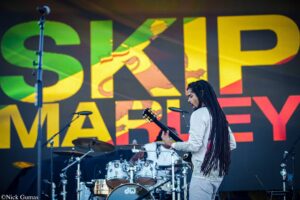 Guitarist with long dreadlocks performing on stage in front of a bright, colorful backdrop and drums visible behind.