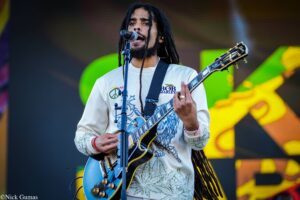 Singer-guitarist with long dreadlocks performing on stage, wearing a light shirt and playing a blue electric guitar in front of a colorful backdrop of lights and banners.