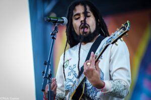 Male guitarist and vocalist with dreadlocks performs on stage, singing into a microphone while playing an electric guitar with a colorful backdrop behind him.