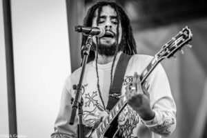 Black-and-white photo of a guitarist singing into a microphone on stage, with long dreadlocks and a floral-patterned guitar design