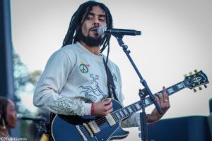Musician with dreadlocks singing into a microphone while playing a dark blue electric guitar on stage outside.