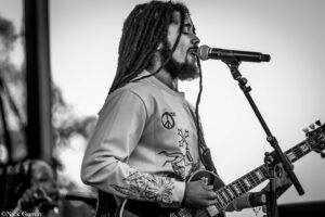 Male guitarist with long dreadlocks singing into a microphone while playing an electric guitar on stage (black-and-white photo).