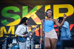 Band performs on a bright stage; foreground singer at mic with guitarist to the left and colorful backdrop behind them.