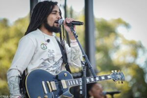 Male musician with long dreadlocks singing into a microphone while playing a black electric guitar on an outdoor stage.
