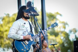 Male guitarist with long dreadlocks performing on an outdoor stage, singing into a microphone.