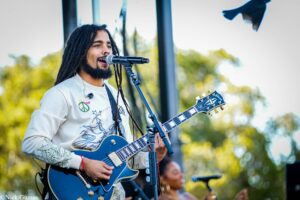 Musician with dreadlocks singing into a microphone while playing a blue electric guitar on an outdoor stage.