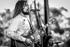 Black-and-white photo of a guitarist with dreadlocks singing into a microphone on stage with an electric guitar in hand for a live performance.