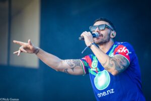 Male singer wearing oversized sunglasses, singing into a microphone with his arm extended on stage.