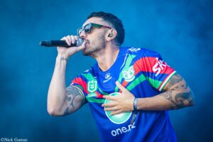 Male singer wearing a colorful blue-green-red jersey and sunglasses, singing into a handheld microphone on stage with visible tattoos on his arm.