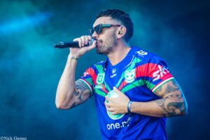 Male singer in a colorful jersey performs on stage, holding a microphone and wearing sunglasses with tattoos visible on his arms.