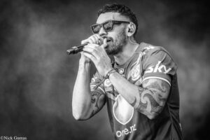 Male singer performing on stage, holding a microphone and wearing sunglasses with visible tattoos on his arm (black-and-white).