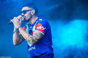 Male singer wearing sunglasses and a blue jersey sings into a microphone on a blue-lit stage, showing tattooed arms and an ear monitor.