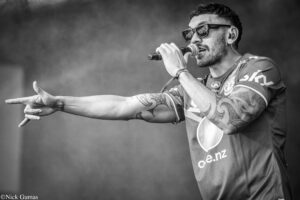 Male singer wearing sunglasses and tattoos, holding a microphone and extending his arm while performing on stage (black and white).