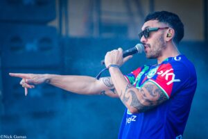 Male singer wearing sunglasses and a blue jersey, microphone in hand, mid-performance on stage with tattoos visible on his arm