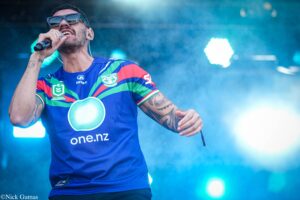 Male singer on stage, wearing sunglasses and a colorful Jersey, singing into a microphone with blue stage lights in the background.
