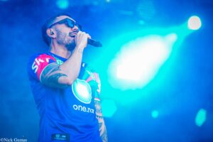 Male singer on stage wearing sunglasses and a blue jersey, gripping a microphone under bright blue stage lights.