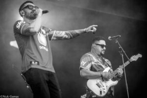Two musicians performing on stage: a tattooed singer wearing sunglasses and pointing, while a guitarist plays nearby in a white electric guitar (black-and-white photo).