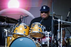 Drummer wearing a navy NY cap and sunglasses behind a gold drum kit on stage with pink light in the background