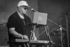 Male musician on stage wearing a white cap and sunglasses, playing a keyboard with a laptop on a stand nearby and a microphone in front of him.
