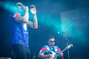 Male singer wearing sunglasses and a blue jersey performs into a microphone on stage, tattooed forearms visible under blue stage lighting.